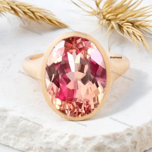 May include: A gold ring with a large oval-shaped pink gemstone. The gemstone has a faceted cut and is set in a bezel setting. The ring is on a white surface.