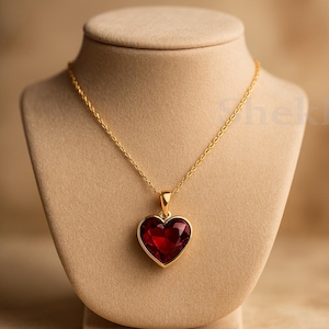 Può includere: Una collana color oro con un ciondolo a forma di cuore rosso intenso. Il ciondolo è incastonato in una cornice dorata e pende da una delicata catena d'oro. La collana è esposta su un busto di gioielleria beige.