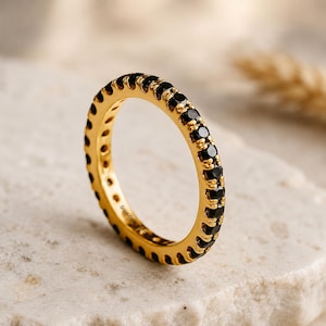 May include: A gold-colored ring with a band of small, black gemstones. The ring is set against a neutral background, highlighting its polished finish and the contrast between the gold and black stones. The ring is a piece of jewelry.