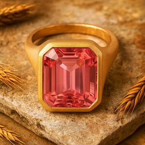 Op de afbeelding: Een gouden ring met een grote, rechthoekige, roze edelsteen. De ring ligt op een getextureerd, lichtbruin oppervlak, met tarwearen op de achtergrond. De edelsteen is veelzijdig.