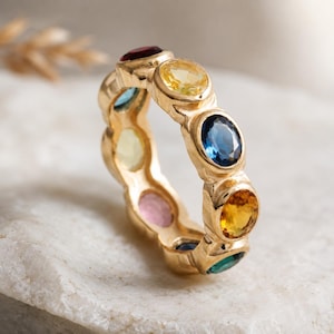 Könnte beinhalten: Ein goldener Ring mit einer Reihe farbiger, ovaler Edelsteine. Die Edelsteine sind rot, gelb, blau und grün und in einem durchgehenden Band angeordnet. Der Ring wird auf einer weißen Oberfläche präsentiert.