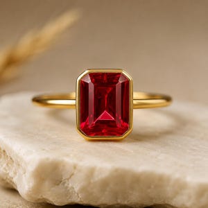 May include: A gold ring featuring a large, rectangular, vibrant red gemstone. The gemstone is set in a gold bezel, and the ring band is a simple, polished gold. The ring is displayed on a light-colored, textured surface.
