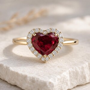 Può includere: Un anello d'oro con una pietra preziosa rosso scuro a forma di cuore, circondata da un alone di piccoli diamanti trasparenti. La fascia dell'anello è in oro liscio. L'anello è appoggiato su una superficie chiara e strutturata.