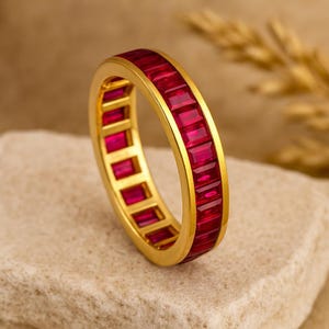 May include: A gold ring with a band of rectangular, vibrant red gemstones. The ring is set on a neutral-toned surface, showcasing the jewelry's design and color. The gemstones are flush with the gold band.
