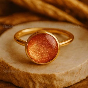 May include: A gold-colored ring with a round, orange-colored stone. The stone has a speckled appearance. The ring is set on a light-colored, textured surface. The brand name "Shekhawatjewels" is visible in the background.