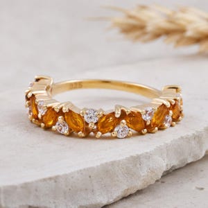 May include: A gold ring featuring a band of orange marquise-cut gemstones and small, round white gemstones. The ring is displayed on a light-coloured, textured surface. The ring is a piece of jewellery.