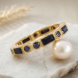 Può includere: Un anello color oro con pietre preziose blu scuro rettangolari e rotonde alternate. L'anello è appoggiato su una superficie di pietra bianca, accanto a una grande perla rotonda bianco sporco. Il bordo dell'anello è decorativo e strutturato.