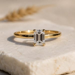 May include: A gold ring with a rectangular, emerald-cut diamond. The diamond is set with four prongs. The ring band is a simple, polished gold. The ring is displayed on a white, textured surface.