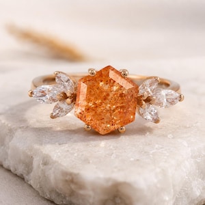 May include: A gold ring featuring a large, orange, hexagonal gemstone as the centerpiece. Flanking the central stone are clusters of white, marquise-cut gemstones. The ring is set on a white marble surface.