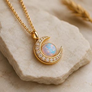 Puede incluir: Un collar dorado con un colgante de media luna. El colgante tiene una esfera central similar a un ópalo y está incrustado con pequeñas piedras preciosas transparentes. El collar descansa sobre una superficie clara y texturizada.