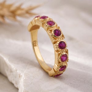 Pode incluir: Um anel dourado com uma série de pedras preciosas redondas rosa-escuro. O anel tem uma banda texturizada e uma moldura decorativa em torno de cada pedra. O anel está sobre uma superfície branca.