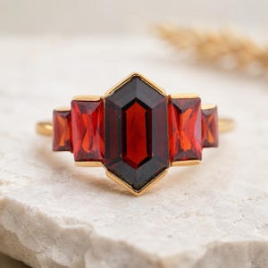 May include: A gold ring featuring a central, dark red, hexagonal gemstone flanked by two rectangular, red gemstones on each side. The ring's band is a thin, polished gold. The gemstones have a faceted cut, reflecting light.
