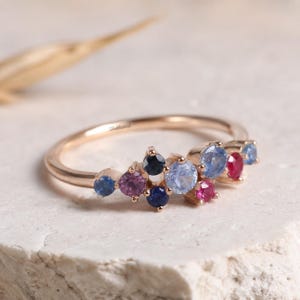 May include: A rose gold ring featuring a cluster of small, round, multi-coloured gemstones including shades of blue, pink, and purple. The band is thin and polished, with the word Shakhawat engraved on the inner surface of the metal ring.