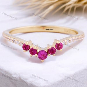 May include: A gold-colored ring with a curved design. The ring features a row of small, clear stones along the top and five bright pink gemstones. The ring is set against a white background.