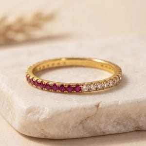 May include: A thin gold band ring featuring a channel setting with a row of round-cut vibrant red rubies along one half and a row of clear, colourless round-cut stones along the other half, creating a distinct two-tone gemstone appearance.