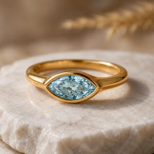 Könnte beinhalten: Ein goldener Ring mit einem hellblauen, marquiseförmigen Edelstein. Der Ring liegt auf einer weißen Marmoroberfläche. Der Edelstein ist der Mittelpunkt, mit einem klaren, facettierten Aussehen. Das Goldband hat eine glatte, polierte Oberfläche.