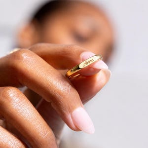 Puede incluir: Un anillo de oro con el nombre "Anna" grabado en el exterior. El anillo es sostenido por una persona con un fondo borroso. El anillo es de diseño de banda simple.