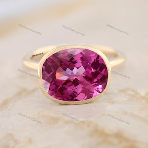Può includere: Un anello d'oro con una pietra ovale rosa. L'anello è su una superficie marrone chiaro.