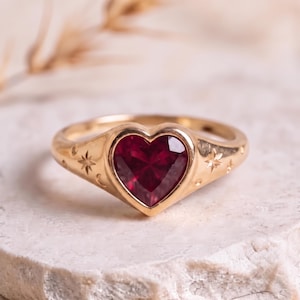 Pode incluir: Um anel de ouro com uma pedra preciosa vermelha escura em forma de coração engastada num bisel. A banda é gravada com pequenos motivos de estrelas e luas crescentes em ambos os lados do engaste, criando um design celestial no metal.