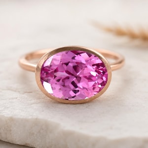 May include: A rose gold ring featuring a large, oval-cut pink gemstone. The gemstone is set in a bezel setting, and the band is a simple, polished design. The ring is displayed on a white surface.