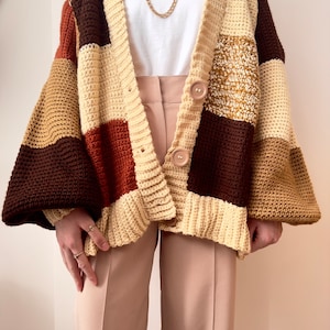 May include: A chunky knit cardigan with a patchwork design in brown, beige, and orange hues. The cardigan has a button closure and long sleeves.