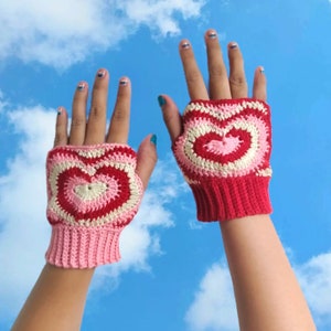 Könnte beinhalten: Ein Paar fingerlose Handschuhe in Pink und Rot mit einem gehäkelten Herzdesign.