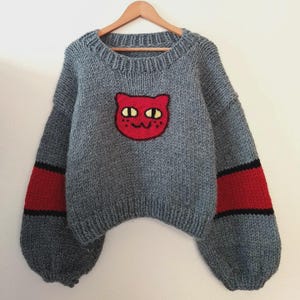 May include: A grey knitted jumper featuring a red cat face motif on the front. The sleeves have red and black stripes. The jumper is displayed on a wooden hanger.