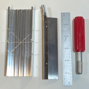 Puede incluir: Una caja de inglete plateada con una sierra plateada y un cuchillo de uso general con mango rojo y hoja plateada. La regla está marcada en pulgadas y tiene el texto "Rule 18" impreso.