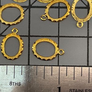 Op de afbeelding: Een verzameling kleine, ovaalvormige goudkleurige metalen bedels met een getextureerde, geschulpte rand en een enkele ring aan de bovenkant. De bedels liggen op een grijze rastermat naast een roestvrijstalen liniaal met maataanduidingen.