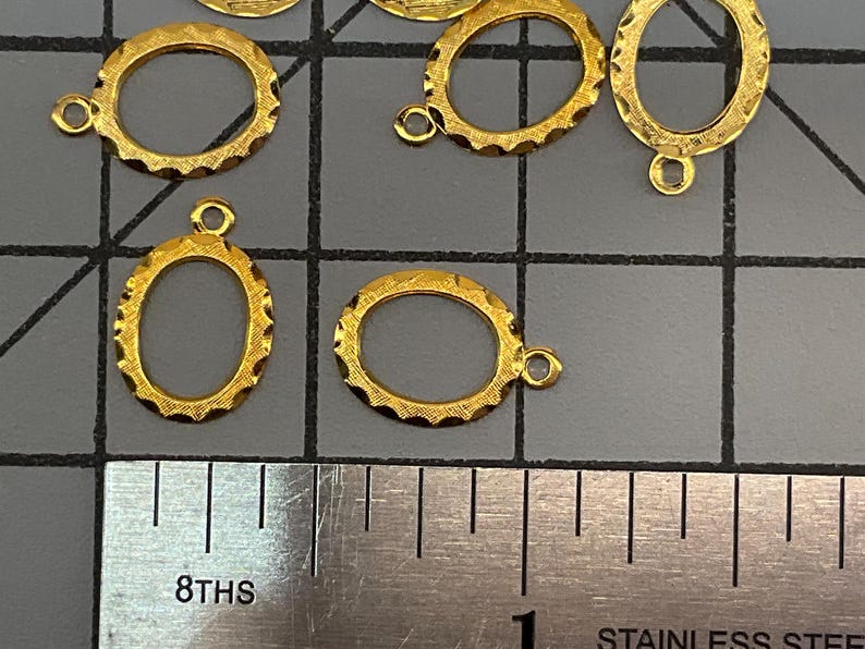 Op de afbeelding: Een verzameling kleine, ovaalvormige goudkleurige metalen bedels met een getextureerde, geschulpte rand en een enkele ring aan de bovenkant. De bedels liggen op een grijze rastermat naast een roestvrijstalen liniaal met maataanduidingen.