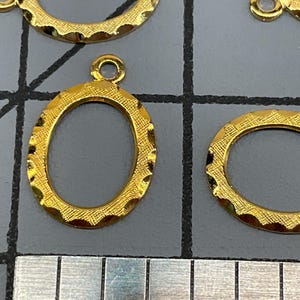 Op de afbeelding: Meerdere goudkleurige ovale metalen bedels met een getextureerde, geschulpte rand en een klein rond oogje aan de bovenkant voor bevestiging. De bedels zijn gerangschikt op een grijs rasteroppervlak naast een liniaal met centimeters.