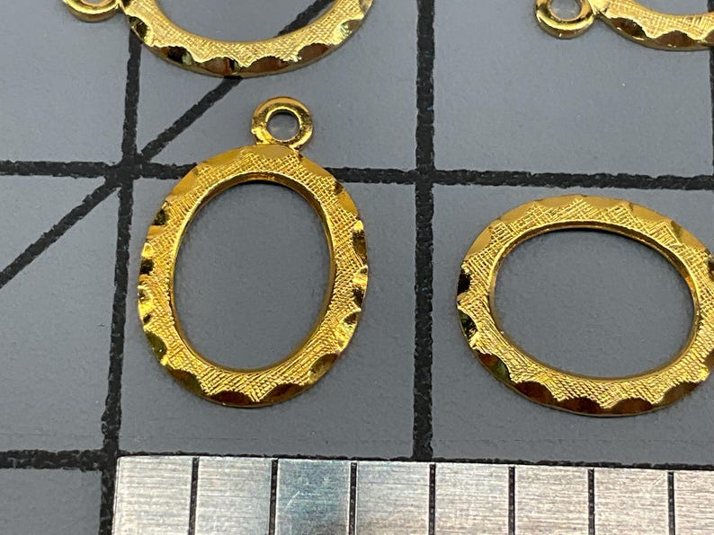 Op de afbeelding: Meerdere goudkleurige ovale metalen bedels met een getextureerde, geschulpte rand en een klein rond oogje aan de bovenkant voor bevestiging. De bedels zijn gerangschikt op een grijs rasteroppervlak naast een liniaal met centimeters.