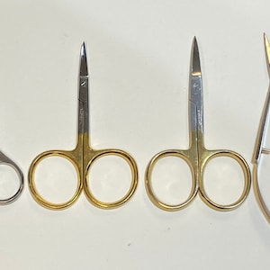 May include: Four pairs of small scissors, two with gold handles and two with silver handles. The scissors are arranged in a row from left to right, with the smallest scissors on the left and the largest scissors on the right. A ruler is visible in the background.