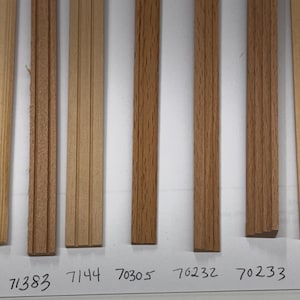 Puede incluir: Una colección de ocho muestras de molduras de madera en varios tonos de marrón y beige. Las muestras están dispuestas verticalmente, mostrando diferentes vetas y acabados de madera. Cada muestra tiene una textura y color únicos, con números escritos debajo.