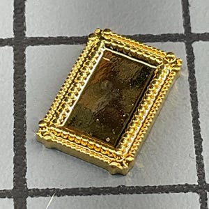 May include: A small rectangular gold-toned metal frame featuring a beaded border design. The center of the frame is filled with a smooth, dark, reflective surface. The object is positioned at an angle, showcasing its metallic finish and texture.