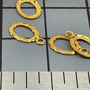 Op de afbeelding: Meerdere goudkleurige ovale sieradenbedels met een getextureerde, gehamerde afwerking. Elke bedel is voorzien van een klein rond oogje aan de bovenkant voor bevestiging. De bedels liggen op een snijmat met een liniaal met cm-aanduiding.