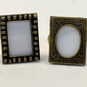 May include: Two antique-style cufflinks. One cufflink has a rectangular frame with a dotted border, the other has an oval frame. Both have a gold-colored metal finish and a white center.