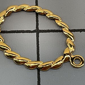 Op de afbeelding: Een goudkleurige metalen sieraadverbinder met een gedraaide, touwachtige textuur in een ovale vorm. Het onderdeel bevat een klein cirkelvormig oogje aan de zijkant voor het verbinden van andere items, gepresenteerd tegen een rasterachtergrond.