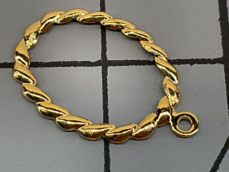 Op de afbeelding: Een goudkleurige metalen sieraadverbinder met een gedraaide, touwachtige textuur in een ovale vorm. Het onderdeel bevat een klein cirkelvormig oogje aan de zijkant voor het verbinden van andere items, gepresenteerd tegen een rasterachtergrond.