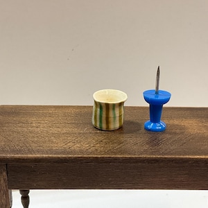 May include: A miniature scene featuring a small, striped ceramic cup and a blue push pin on a wooden table. The cup has green and brown stripes. The push pin has a silver pin and a blue head.