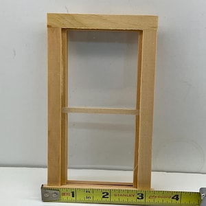 May include: A small, unfinished wooden window frame with a clear pane. The frame is rectangular, with a horizontal divider. A yellow measuring tape shows the window is approximately 7.6 cm wide.