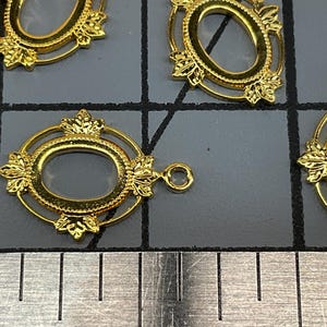 Op de afbeelding: Vier goudkleurige ovale metalen hangers met decoratieve bladachtige accenten aan de boven- en onderkant. Elke hanger heeft een centrale ovale opening en een klein rond oogje aan de rand, geplaatst boven een metalen liniaal voor schaal.