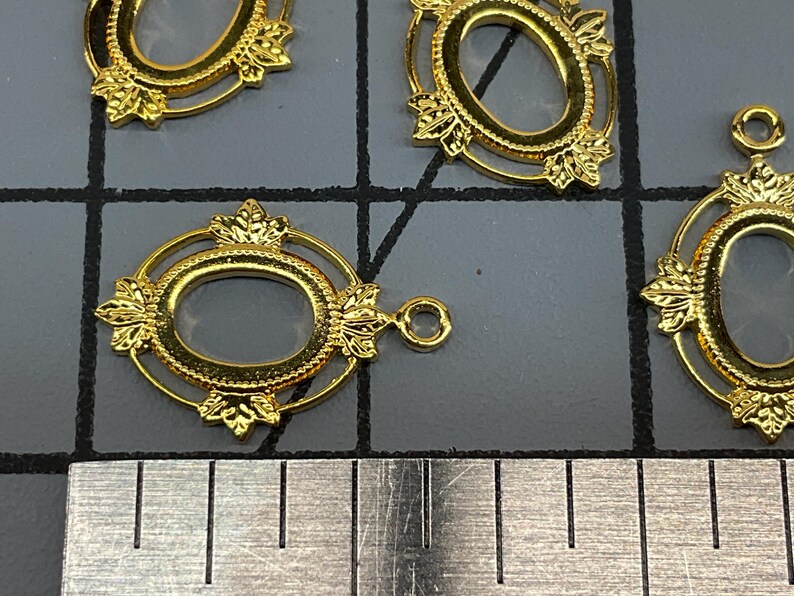 Op de afbeelding: Vier goudkleurige ovale metalen hangers met decoratieve bladachtige accenten aan de boven- en onderkant. Elke hanger heeft een centrale ovale opening en een klein rond oogje aan de rand, geplaatst boven een metalen liniaal voor schaal.