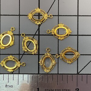 Op de afbeelding: Zeven goudkleurige ovale framebedels met ornamentale details en een klein lusje aan de bovenkant voor bevestiging. De bedels zijn gerangschikt op een rastermat naast een roestvrijstalen liniaal, die een hoogte van ongeveer 1,9 cm aangeeft.