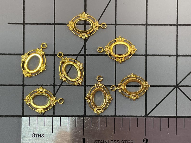 Op de afbeelding: Zeven goudkleurige ovale framebedels met ornamentale details en een klein lusje aan de bovenkant voor bevestiging. De bedels zijn gerangschikt op een rastermat naast een roestvrijstalen liniaal, die een hoogte van ongeveer 1,9 cm aangeeft.