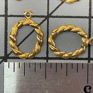 Op de afbeelding: Twee goudkleurige ovale bedels met een gedraaide touwtextuur en een klein ringetje aan de bovenkant. De bedels liggen op een rastermat naast een metalen liniaal, waaruit blijkt dat ze elk ongeveer 1,3 cm hoog en breed zijn.