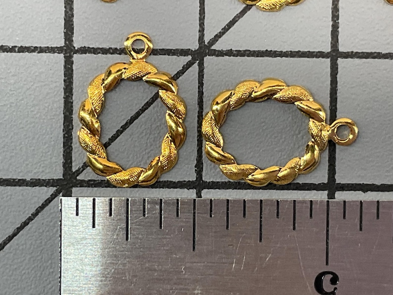 Op de afbeelding: Twee goudkleurige ovale bedels met een gedraaide touwtextuur en een klein ringetje aan de bovenkant. De bedels liggen op een rastermat naast een metalen liniaal, waaruit blijkt dat ze elk ongeveer 1,3 cm hoog en breed zijn.