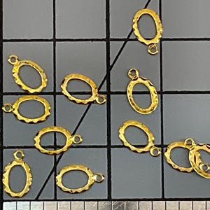May include: Ten small gold-colored oval charms with a textured, scalloped edge and a single jump ring at the top. The charms are arranged on a dark gray cutting mat with a visible grid and a ruler at the bottom edge for scale reference.