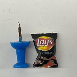 May include: A miniature blue push pin next to a small black bag of Lay's Barbecue potato chips. The chip bag has the Lay's logo in yellow and red, with the word "Barbecue" below.