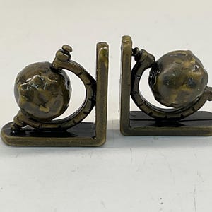 May include: A pair of bronze-colored bookends. Each bookend features a globe design held within a curved frame, resting on a rectangular base. The globes have a textured surface, and the overall design is vintage.
